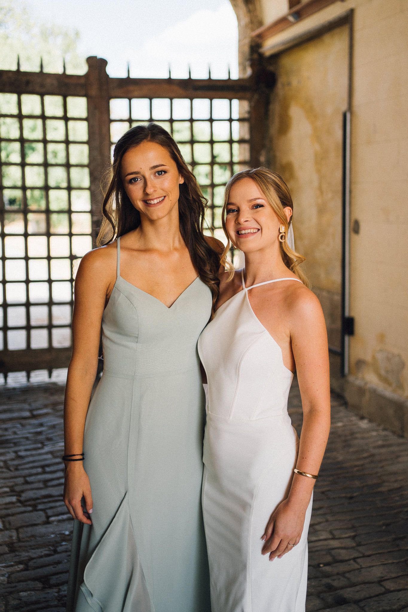 Two women in sage green and white dresses pose together in an elegant venue with natural lighting.
