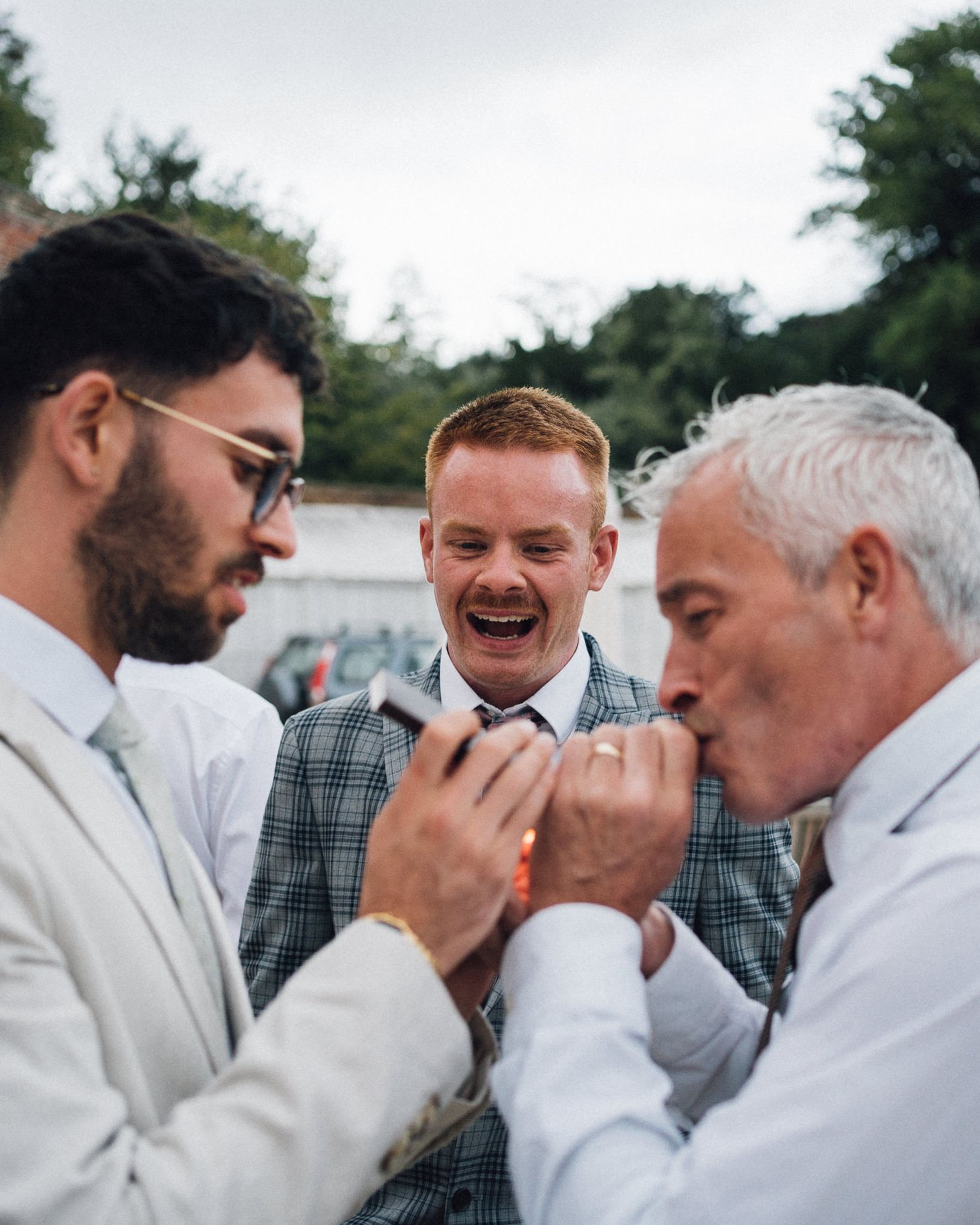 Three wedding guests share a lighthearted moment outdoors, with one wearing a plaid vest between two in light suits.