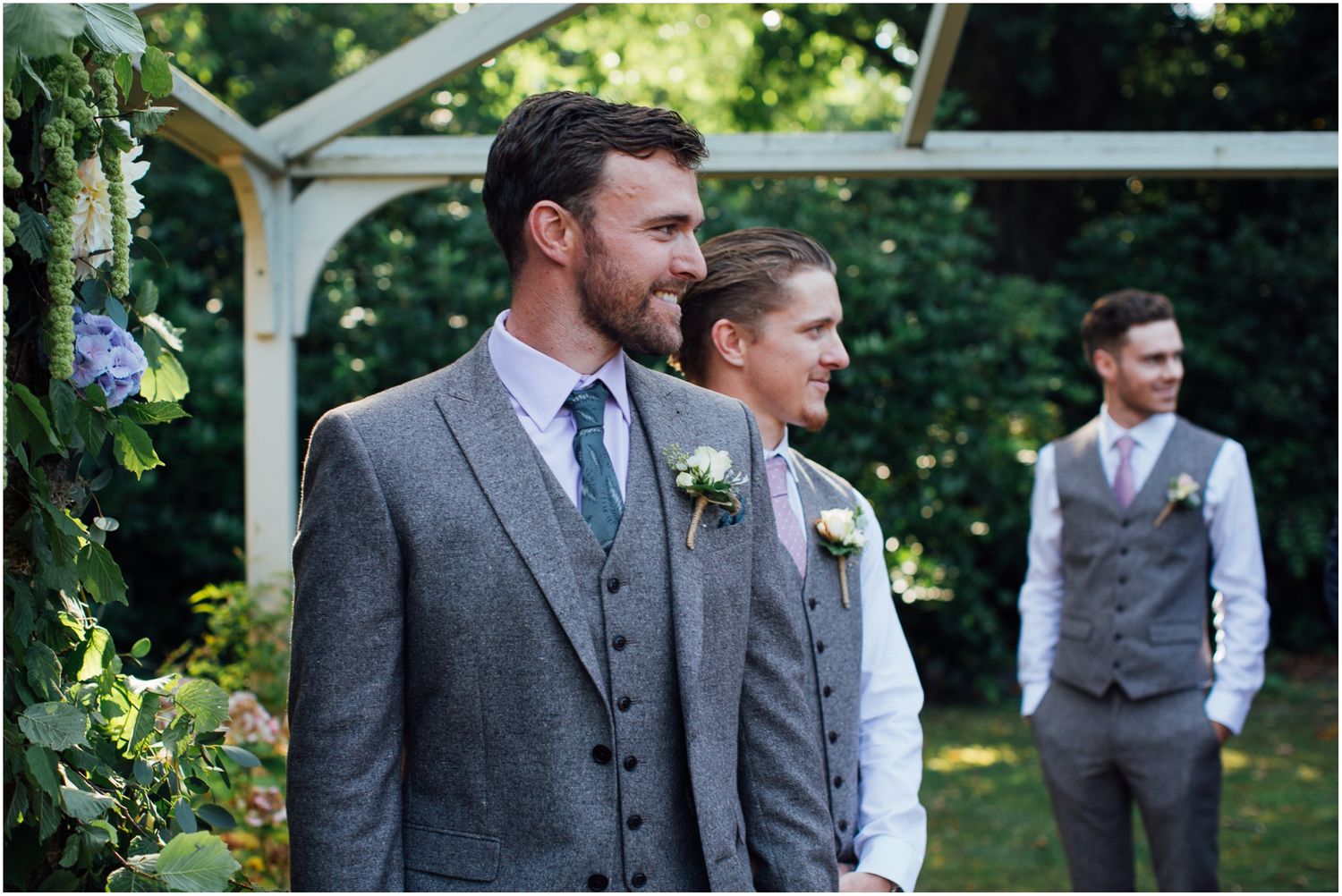 Groomsmen in grey suits standing under garden arbor.