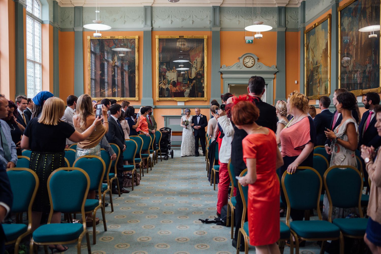 People sit in rows of teal chairs in a long hallway with orange walls and pendant lighting.
