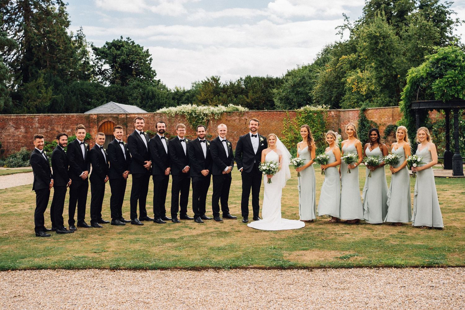 Wedding party lined up outdoors with groomsmen in black suits and bridesmaids in pale yellow dresses by brick wall.