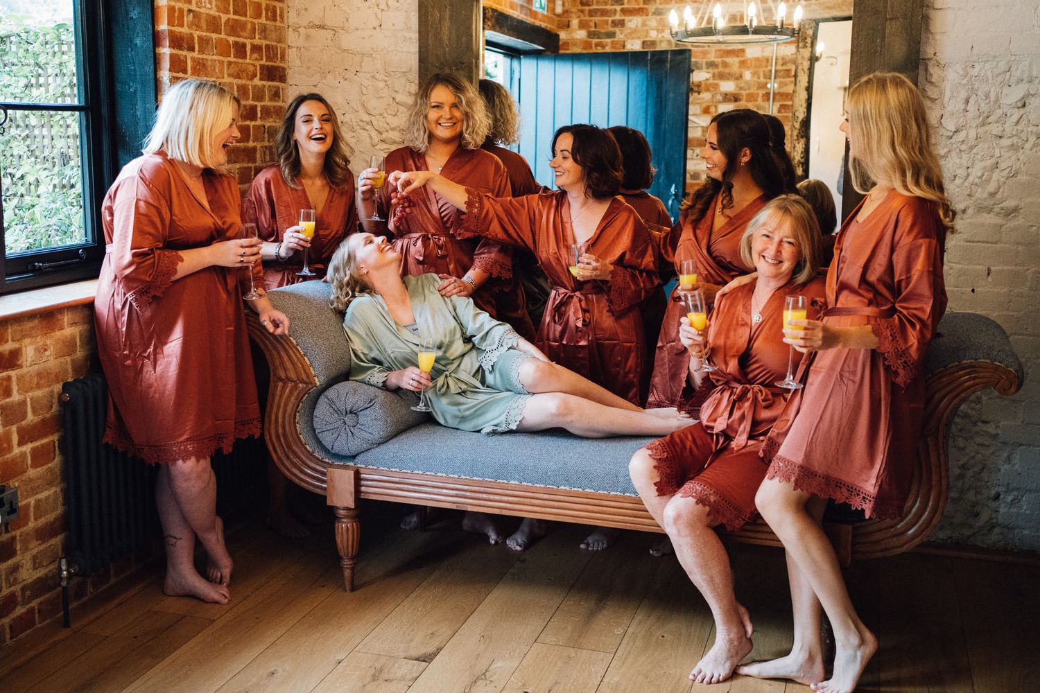 Group of women in silk robes laughing together on a couch in a bridal suite.