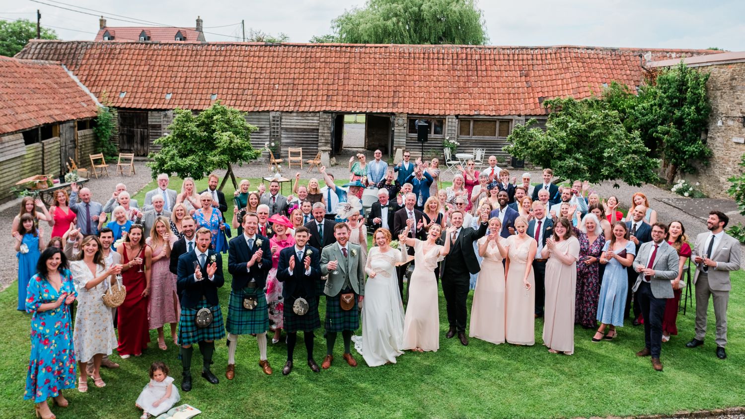 Large wedding party gathering outdoors in front of rustic red-roofed barn with guests celebrating on green lawn.