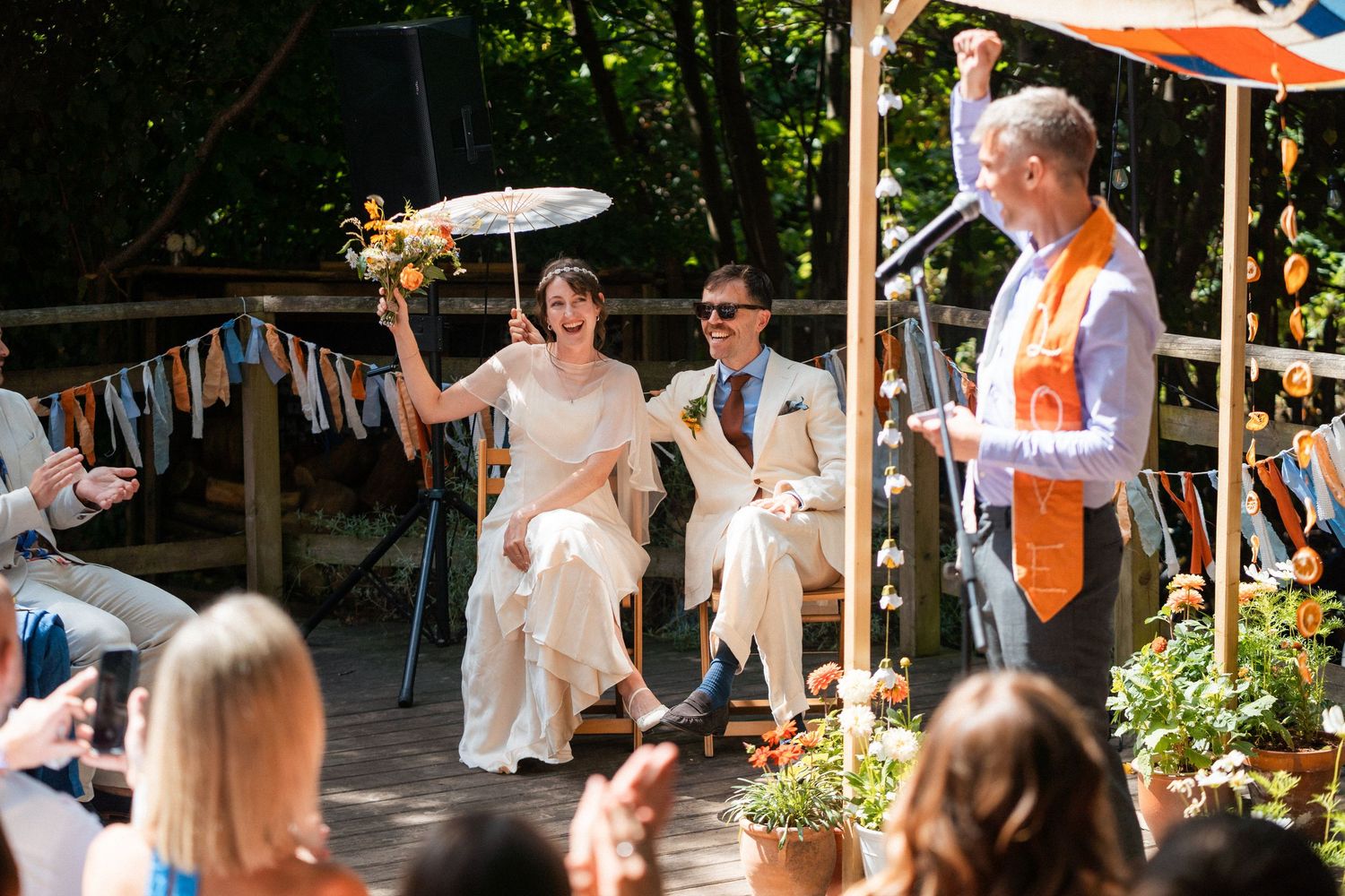 Bride and groom celebrate during outdoor wedding ceremony on wooden deck with colorful bunting decorations.
