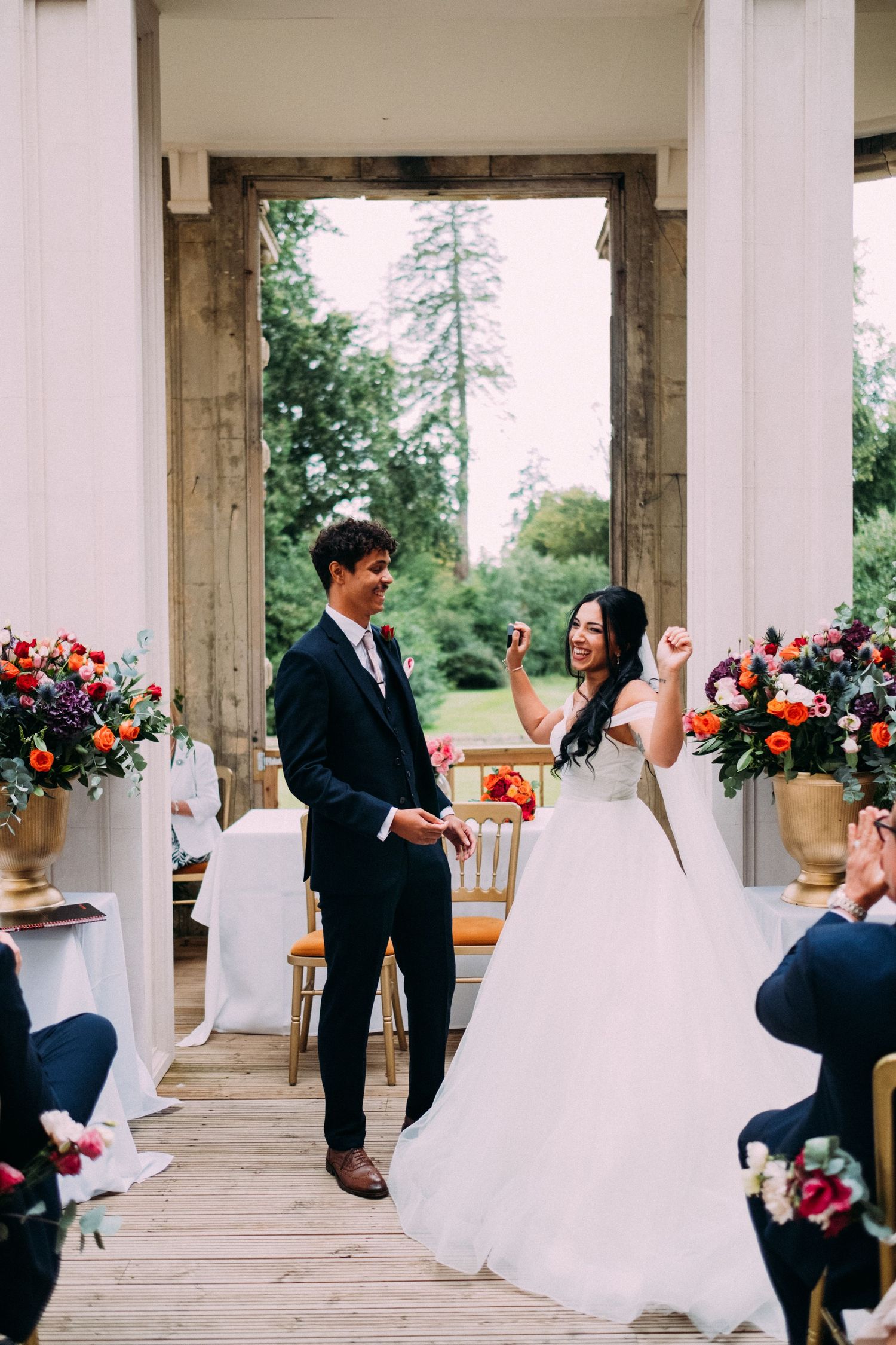 Wedding ceremony setup with white chairs and floral arrangements in an outdoor columned pavilion.
