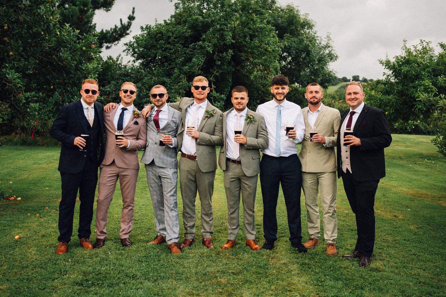 Wedding party in coordinating khaki and dark suits poses together on green lawn during outdoor celebration.