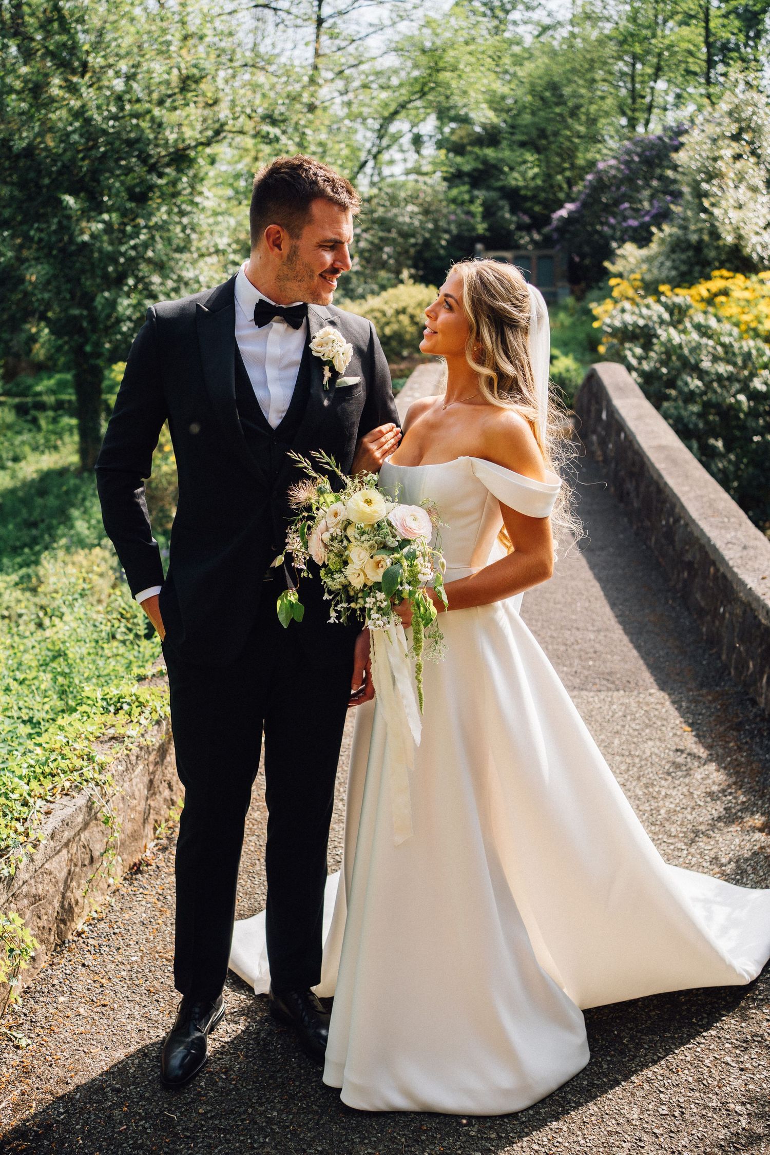 A romantic wedding photoshoot featuring a couple in formal attire on a stone bridge pathway.