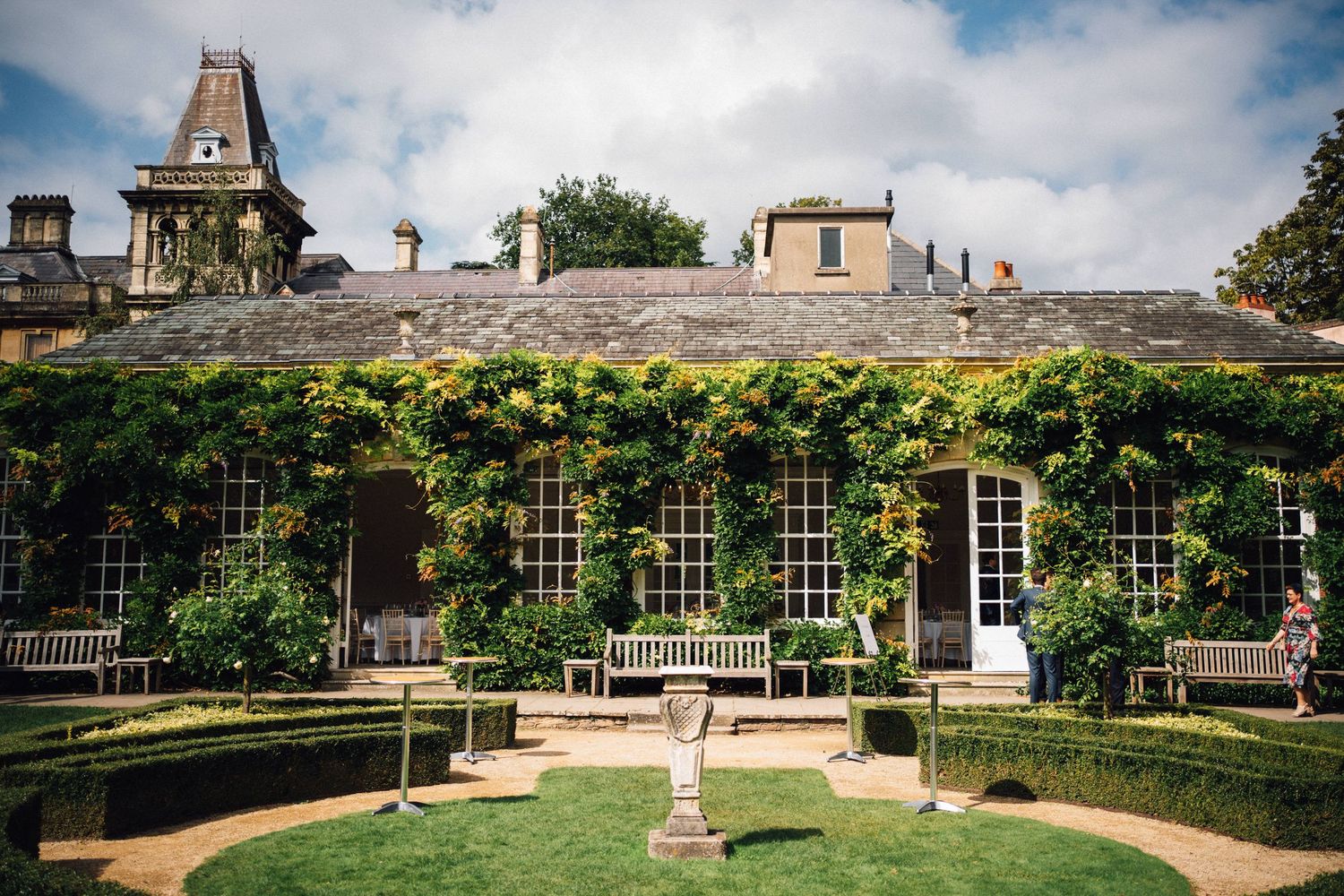 Ivy-covered stone manor house with manicured garden, sundial centerpiece, and benches under cloudy sky.