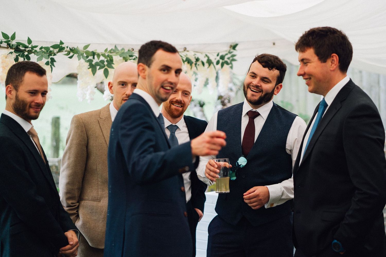 A group of well-dressed men in suits share a laugh during a social gathering with decorative greenery in the background.