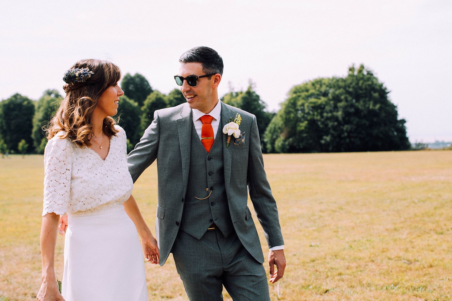 A newlywed couple walks hand in hand across a sunny meadow in elegant wedding attire.