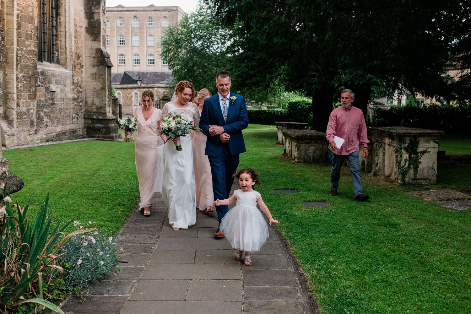 Wedding procession walking along church garden path with small child in white dress.