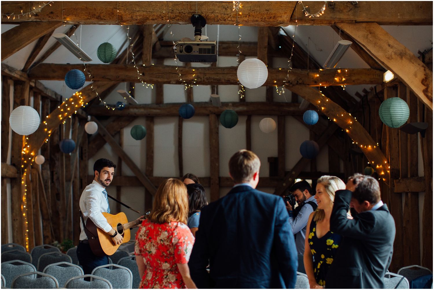 Wedding guests mingling inside a rustic barn venue decorated with string lights and paper lanterns.