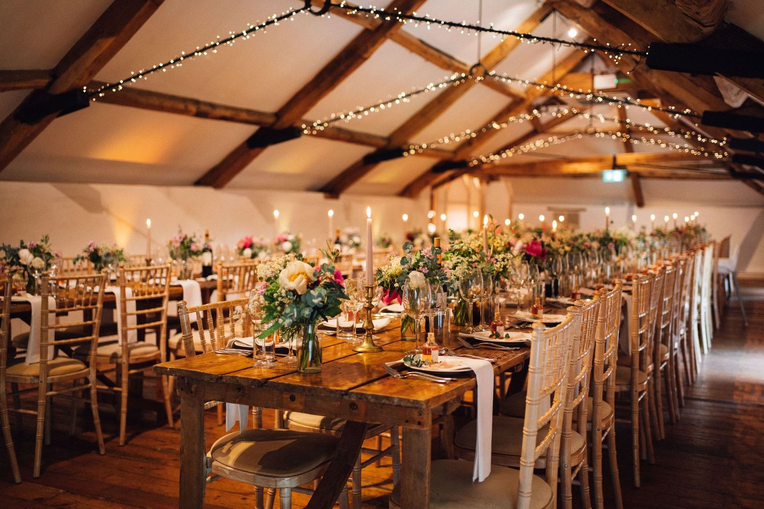 Long banquet tables set up in a rustic barn venue with string lights overhead.