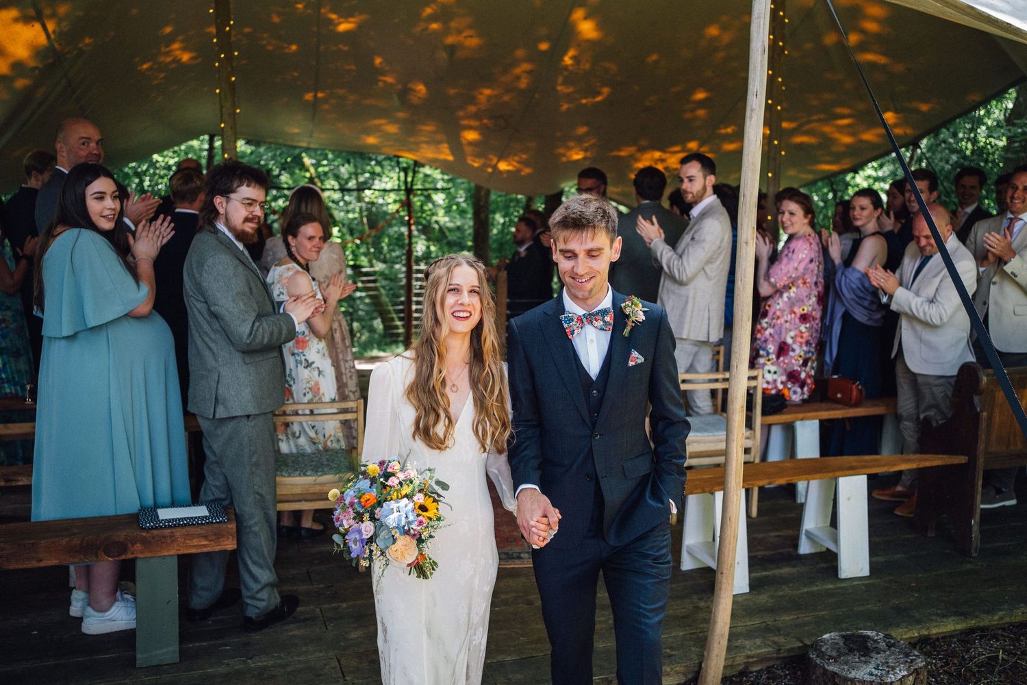 A couple walks down the aisle at an outdoor tent wedding while guests applaud.