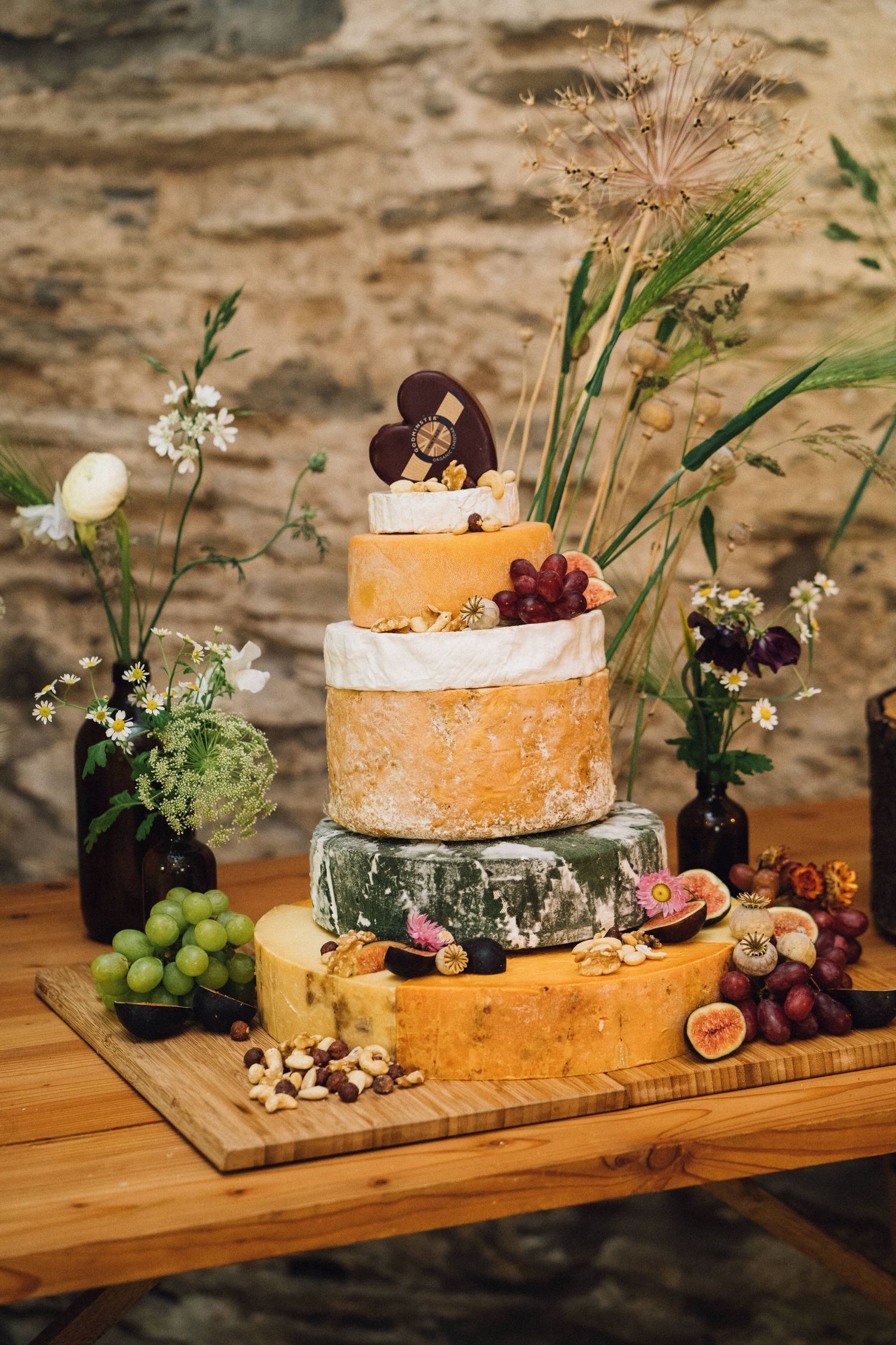 A rustic cheese wheel wedding cake decorated with fresh figs, grapes and flowers on a wooden board.