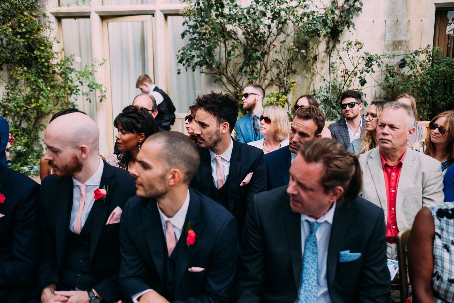 Wedding guests in formal attire seated outdoors at a ceremony venue with greenery in the background.