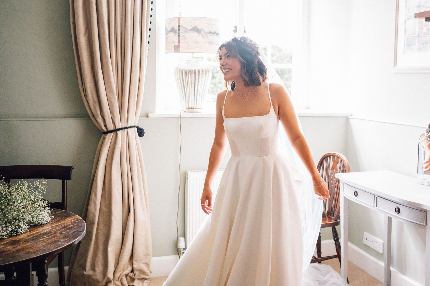 Sunlit bridal preparation scene with flowing white wedding dress near window.