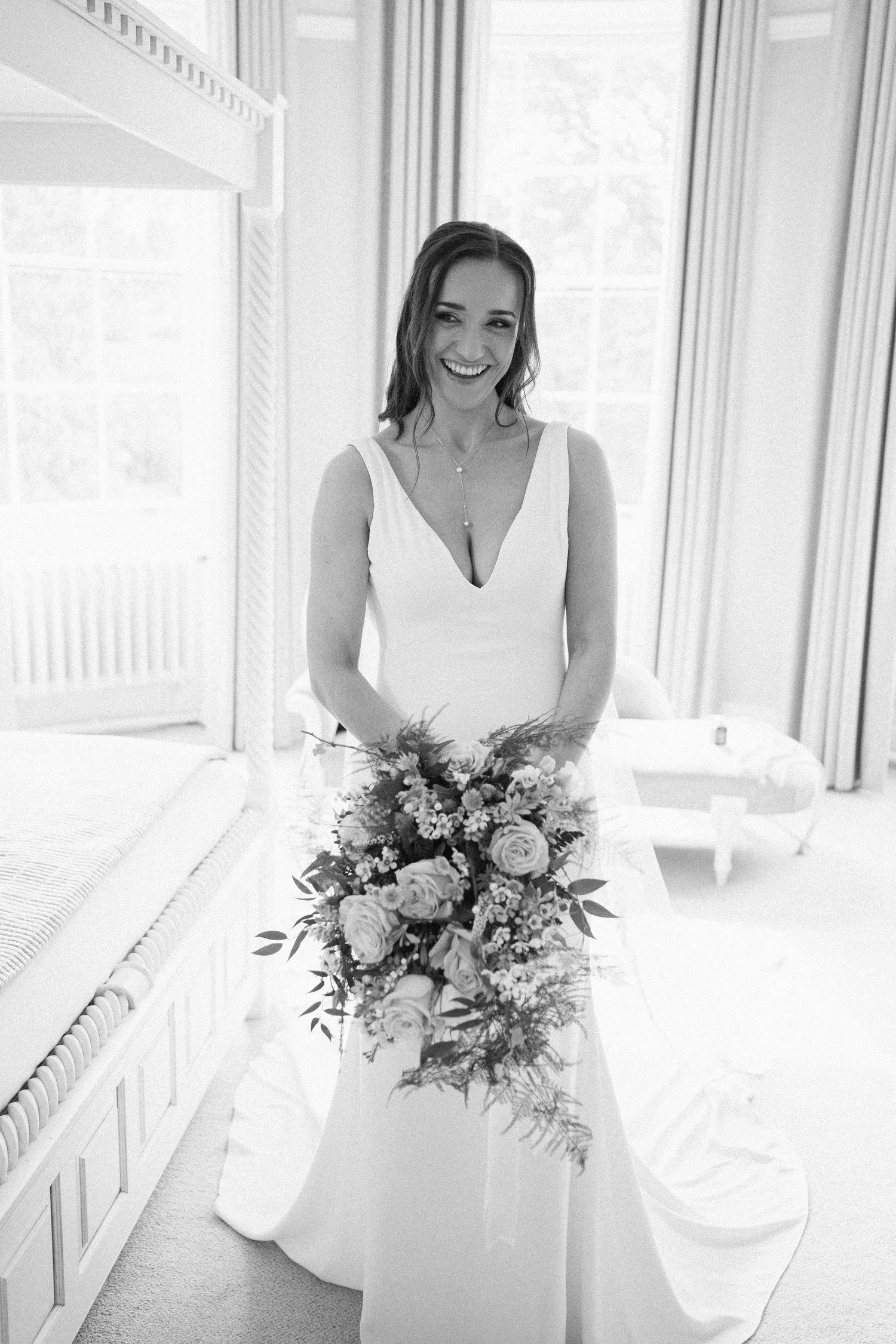 A bride holds a beautiful bouquet of mixed flowers while standing in front of large windows in natural light.