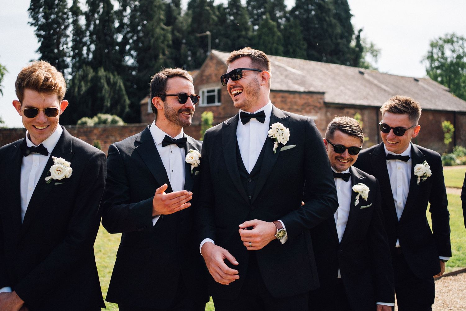 Groomsmen in black tuxedos and sunglasses pose together outdoors for wedding photos.