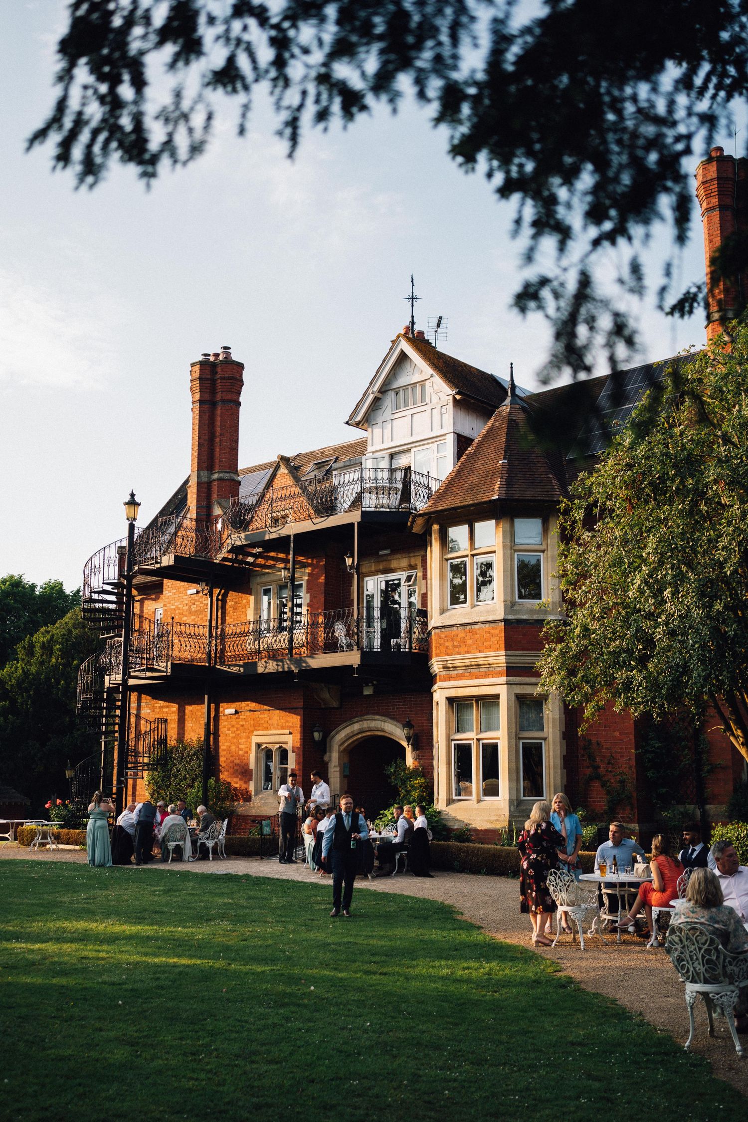 Historic Victorian mansion with red brick and Tudor details serves as a gathering spot with outdoor seating on a sunny lawn.