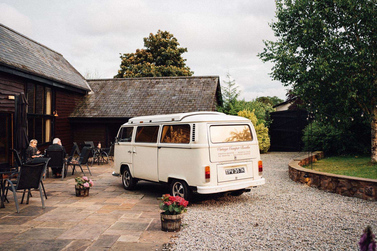 Classic white Volkswagen camper van parked on gravel driveway outside rustic thatched-roof building.