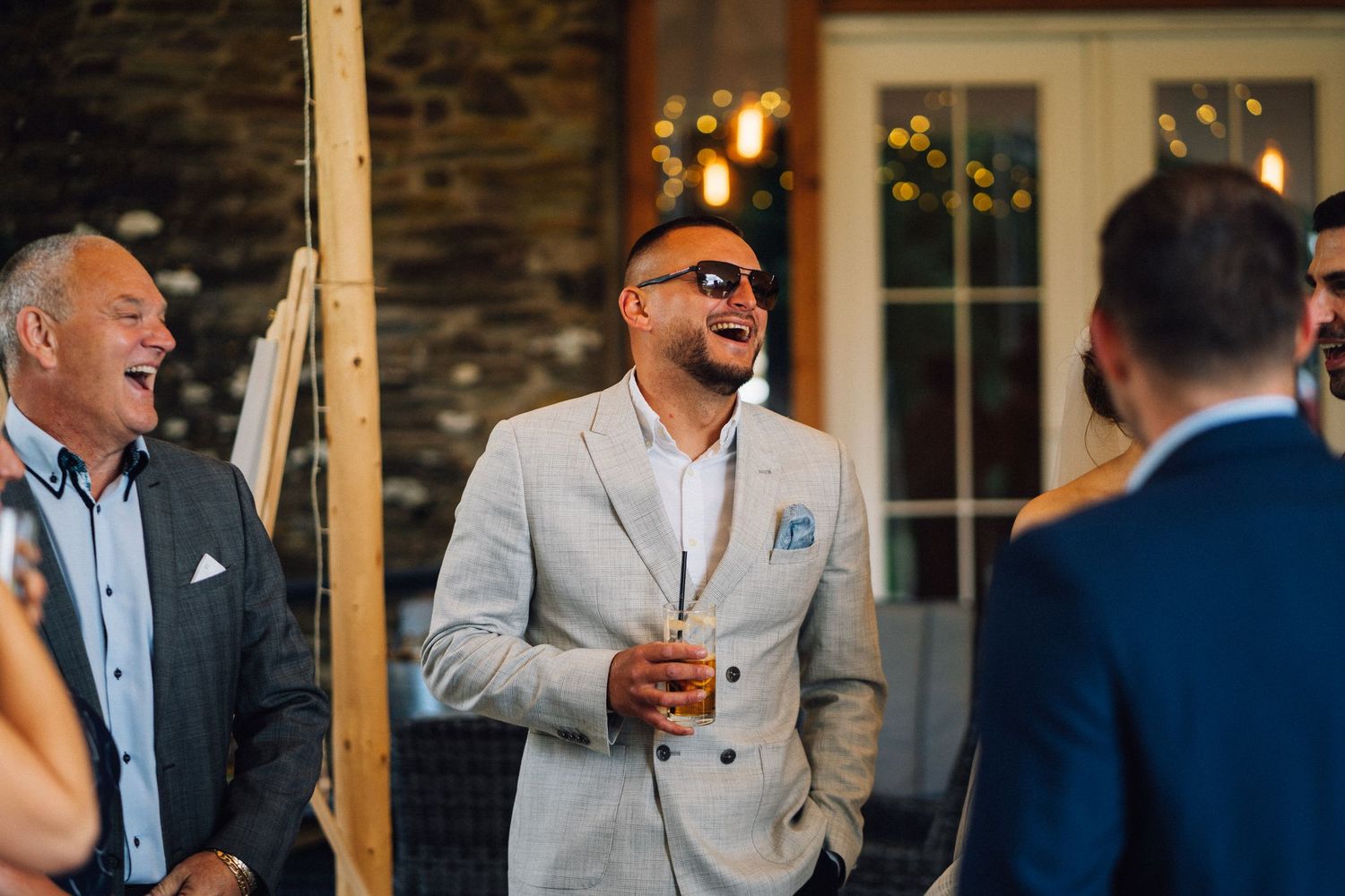 Wedding guests share laughter and drinks during an evening reception at a rustic stone venue.