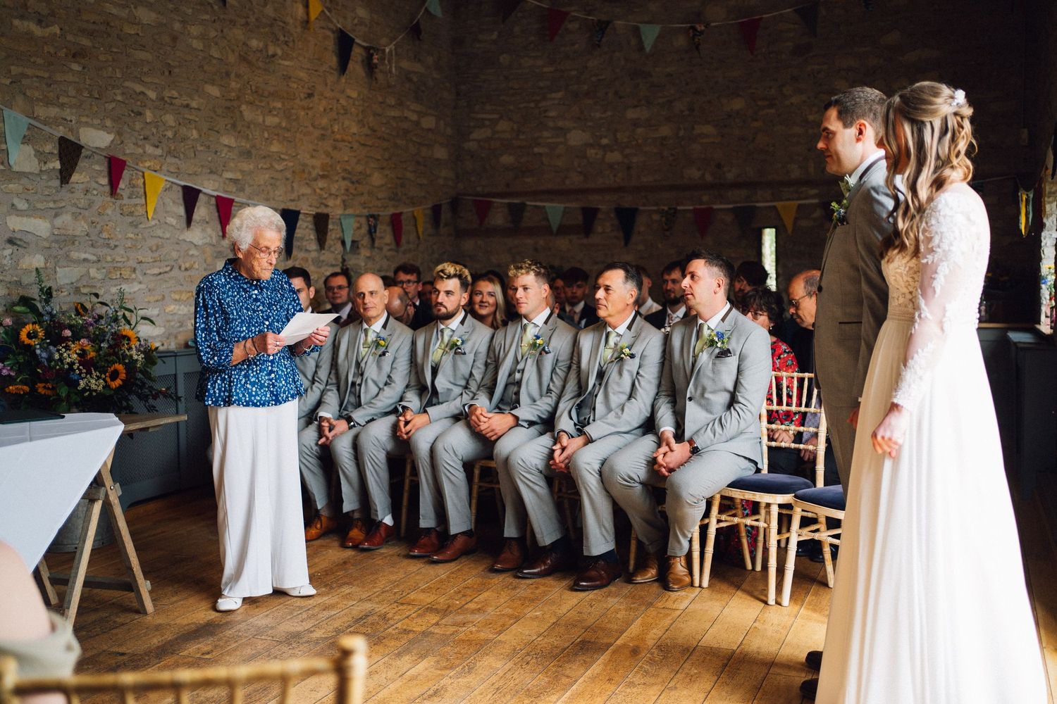 Wedding party members seated in a row along a rustic brick wall with string lights above during celebration.