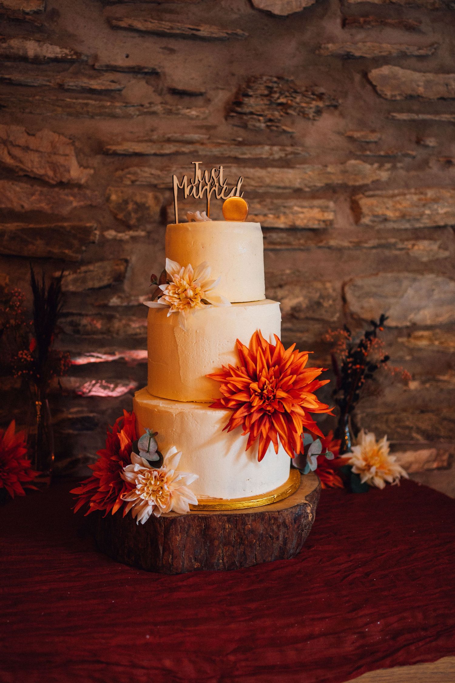 Rustic wedding cake decorated with orange dahlias and autumn flowers displayed on wood slice against stone wall.