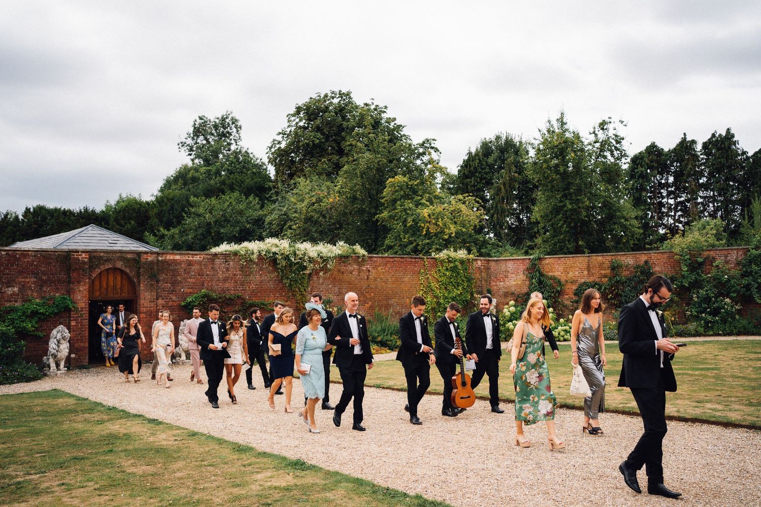 Wedding guests walking in a line along a gravel path beside a rustic brick wall in a garden setting.