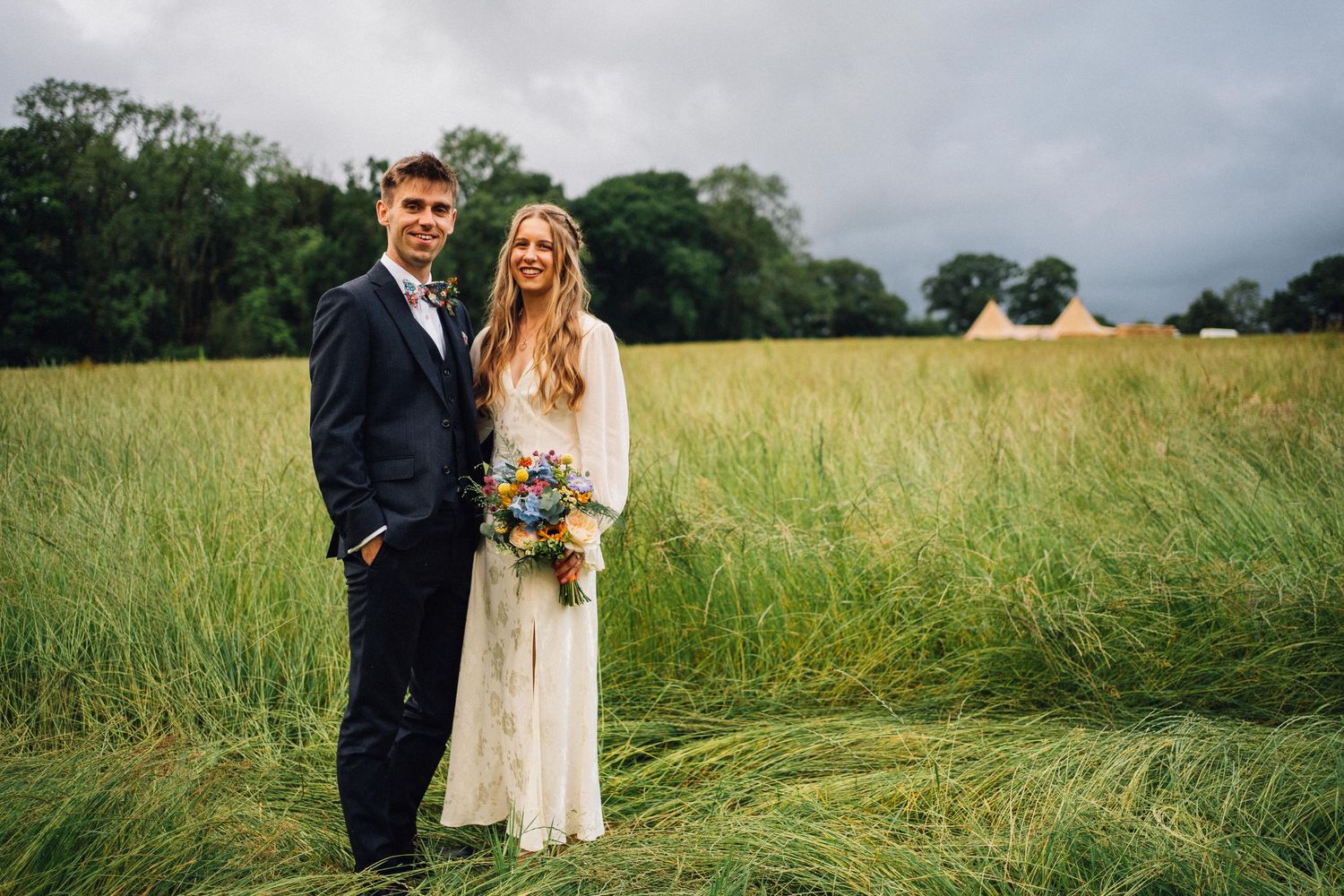 Couple stands together in a lush green field on their wedding day as sunlight filters through the trees.