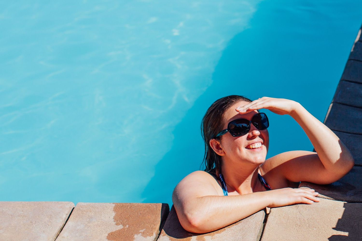 Person relaxing at the edge of a bright blue infinity pool on a sunny day.