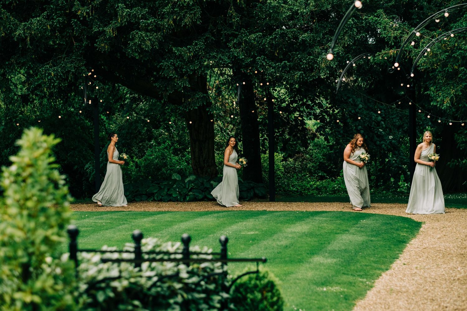 Figures in light gray dresses walk along a curved garden path during an outdoor ceremony.