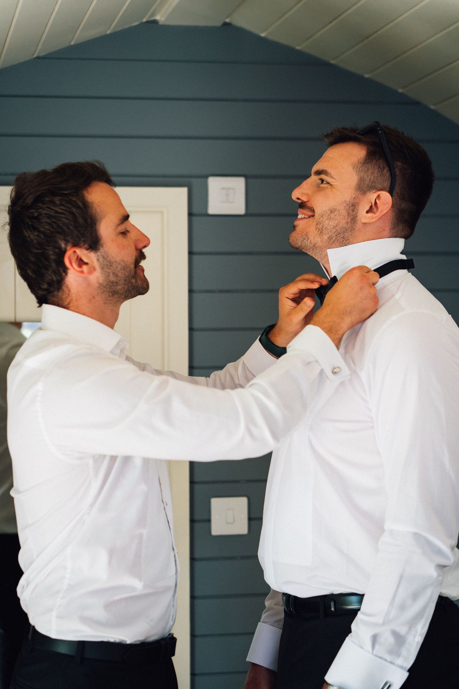 Two people in white dress shirts help each other adjust bow ties and neckwear while getting ready.