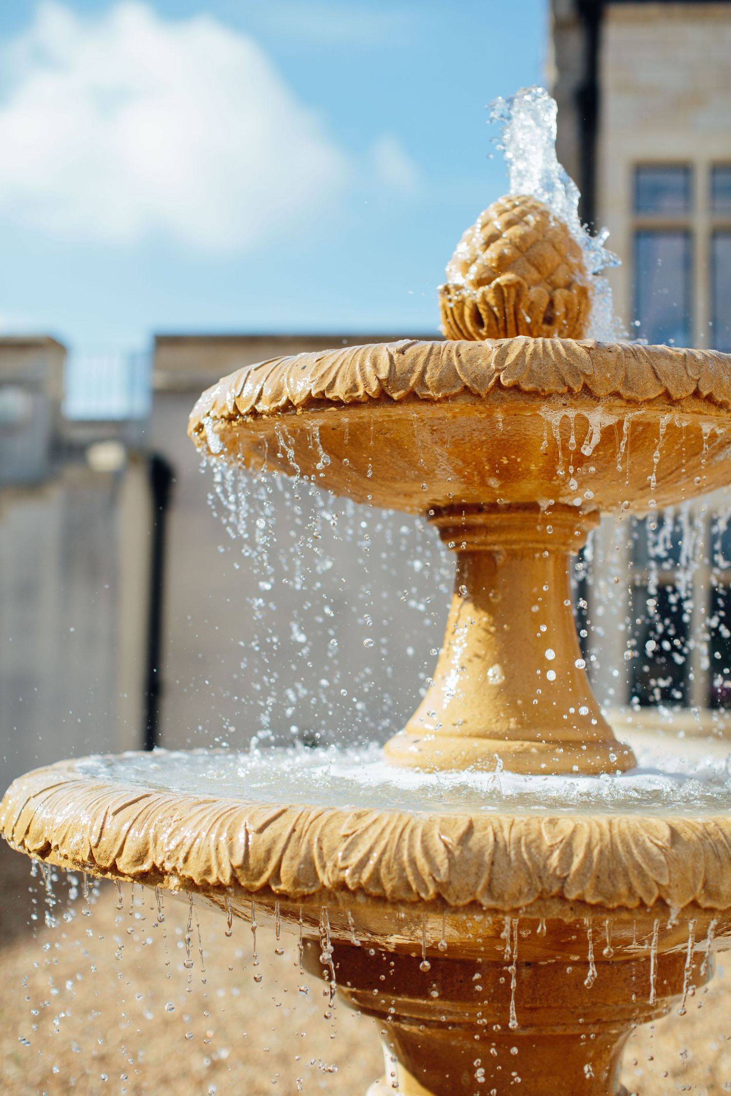 Close-up view of an ornate golden fountain with water droplets spraying in sunlight against a blue sky.