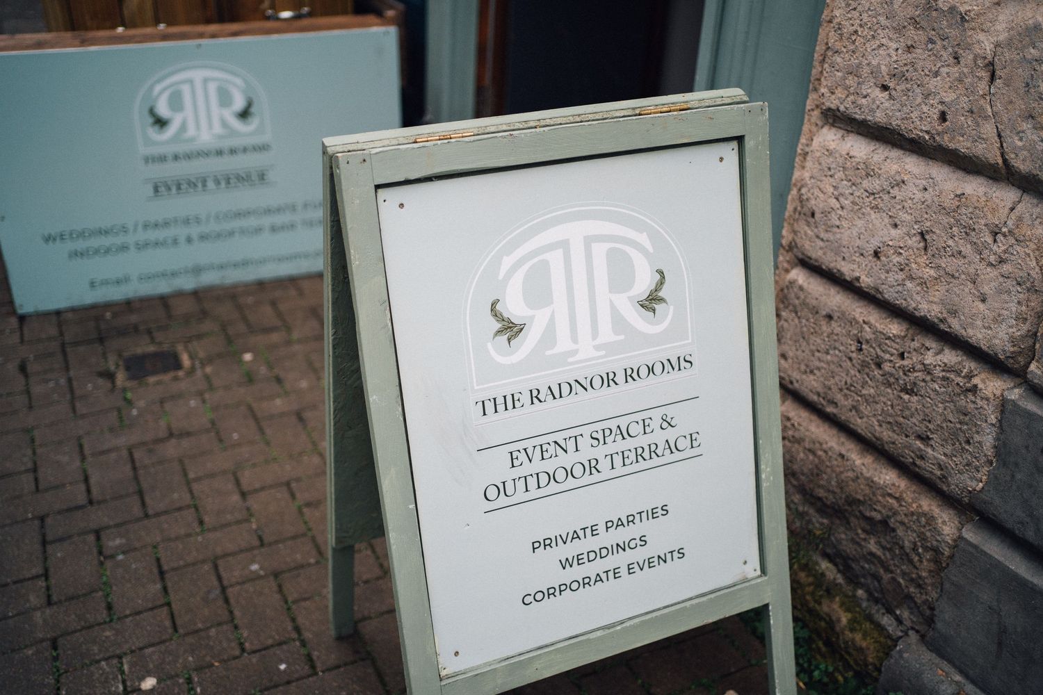 A framed business sign for 'The Raven Room' with an elegant logo design stands on a brick sidewalk.