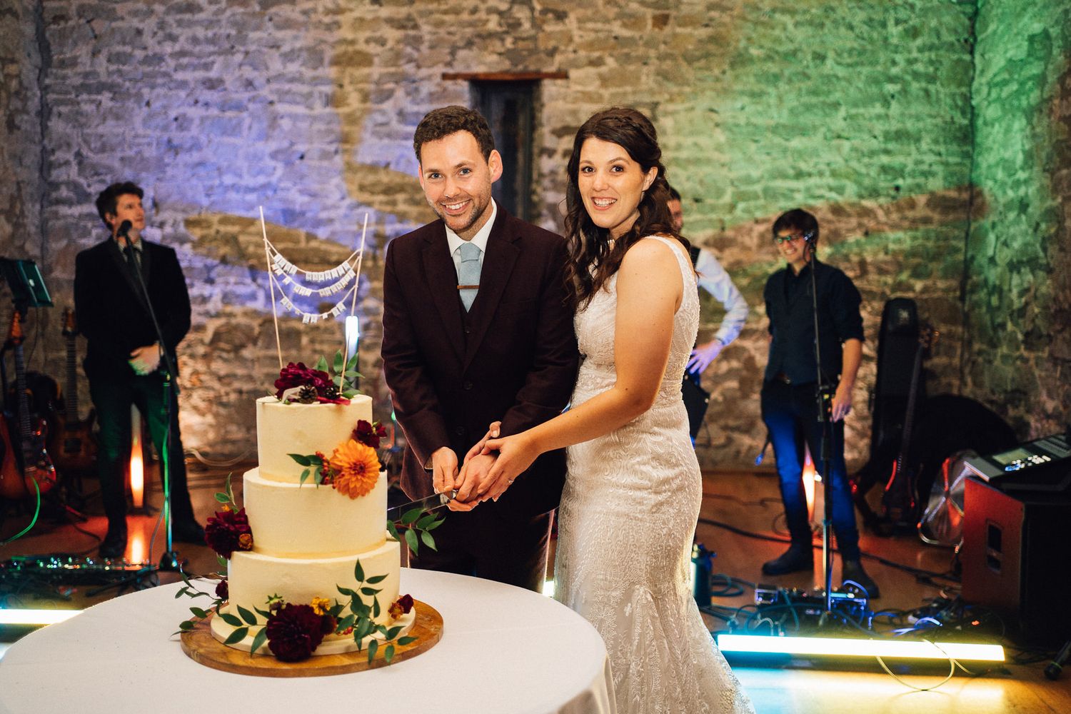 Couple cuts their wedding cake together at a rustic brick venue with colorful lighting in the background.