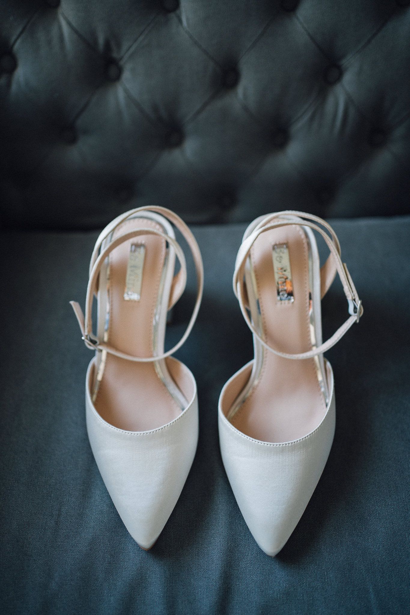 Elegant white pointed-toe bridal shoes with ankle straps displayed on a tufted dark blue velvet surface.