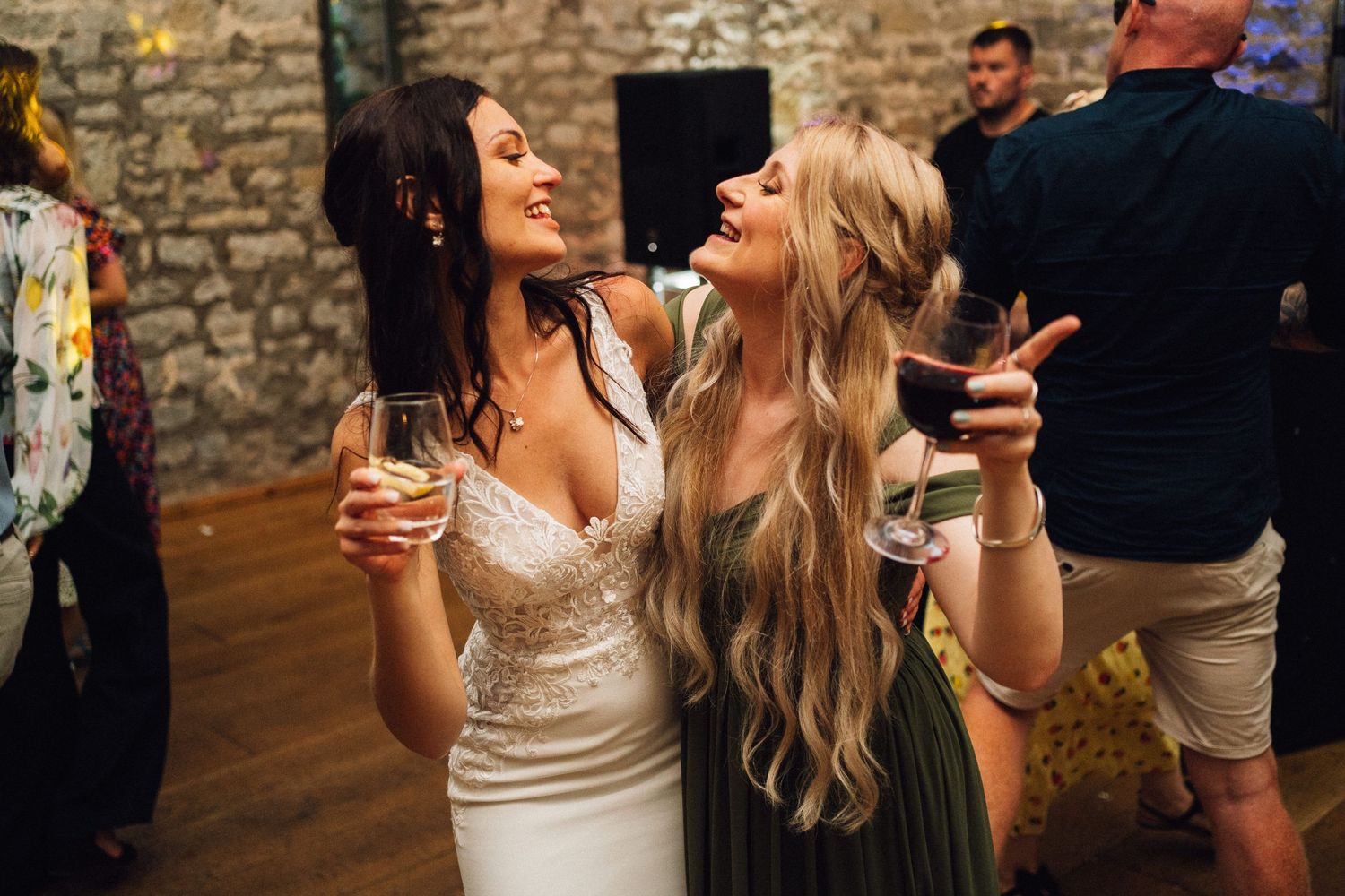 Wedding guests laugh and dance while holding drinks at a rustic stone-walled reception venue.