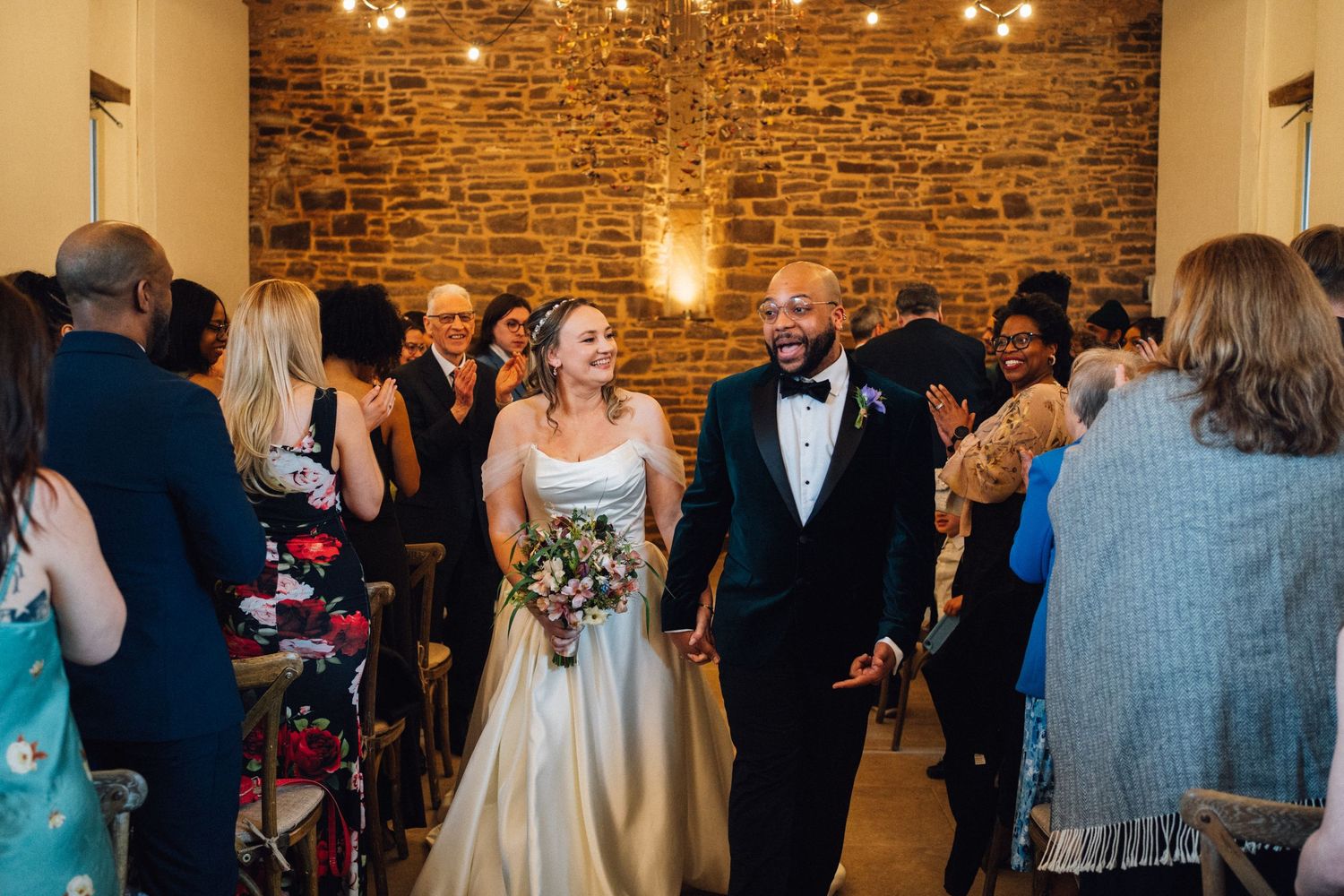 Wedding guests celebrate and dance together in a rustic stone-walled venue with warm lighting.