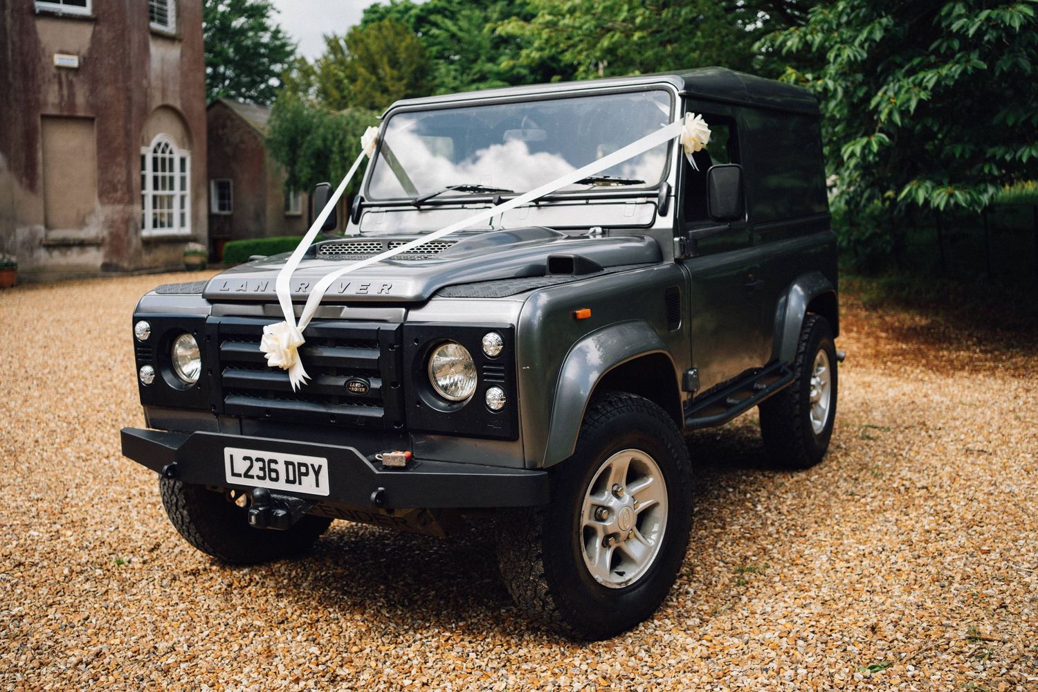 Black Land Rover Defender decorated with wedding ribbons parked on a gravel driveway.