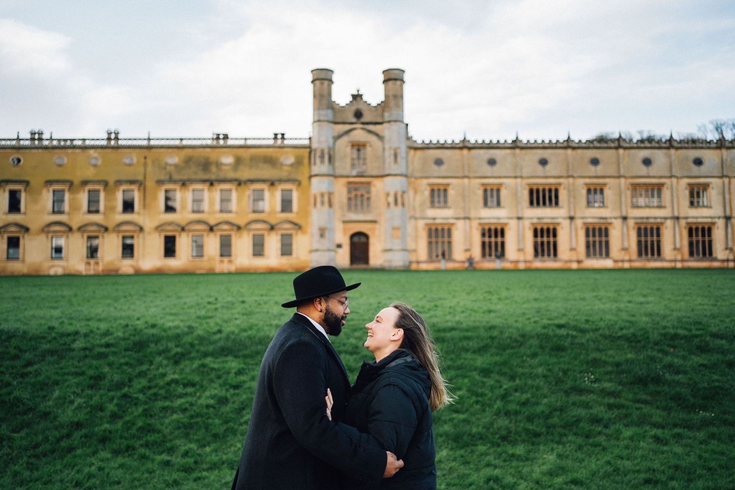 Romantic couple posing in front of historic university building on a grassy lawn.