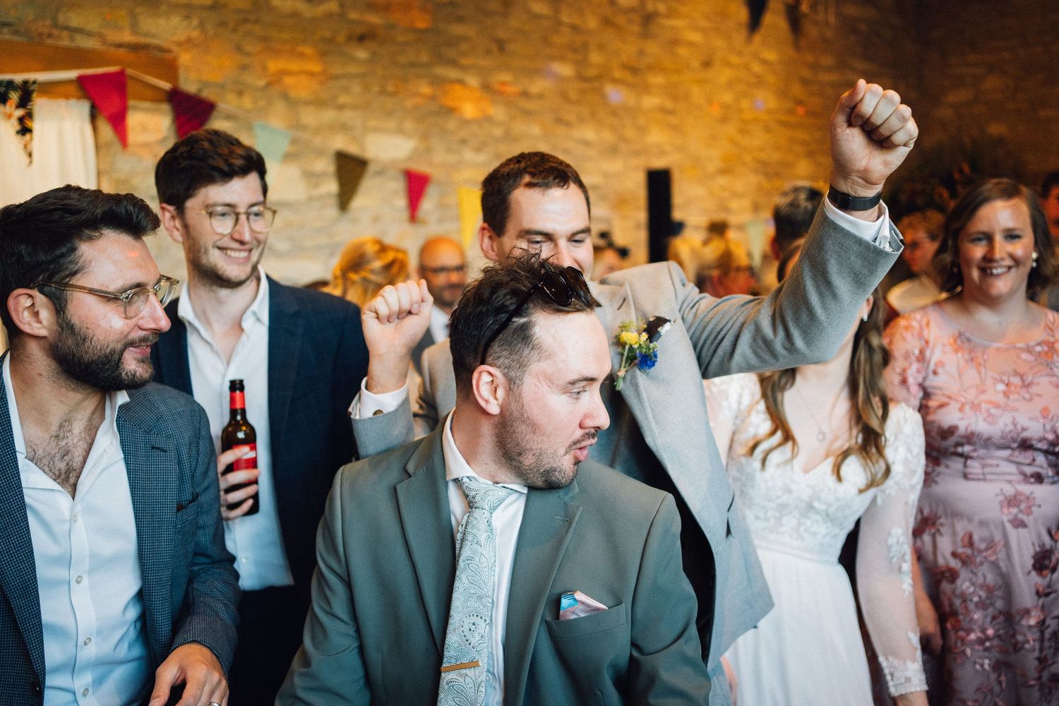 Wedding party celebrating with raised arms in a stone-walled venue with festive lighting.