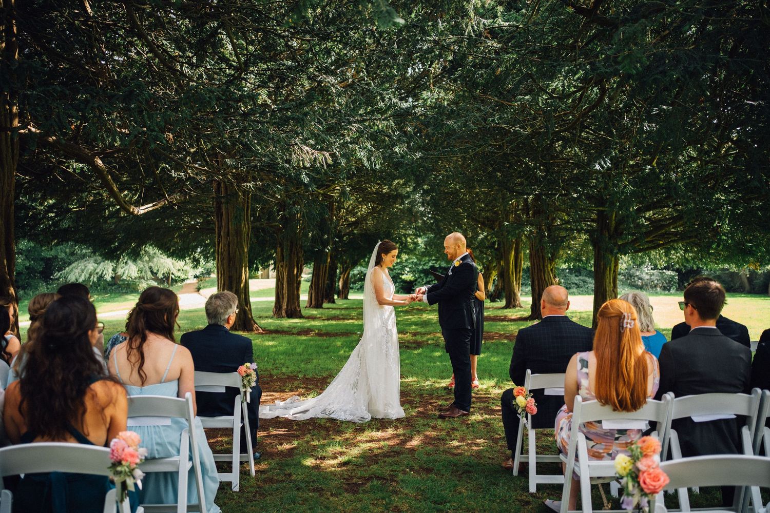 Outdoor wedding ceremony with white chairs arranged in rows under tall trees.