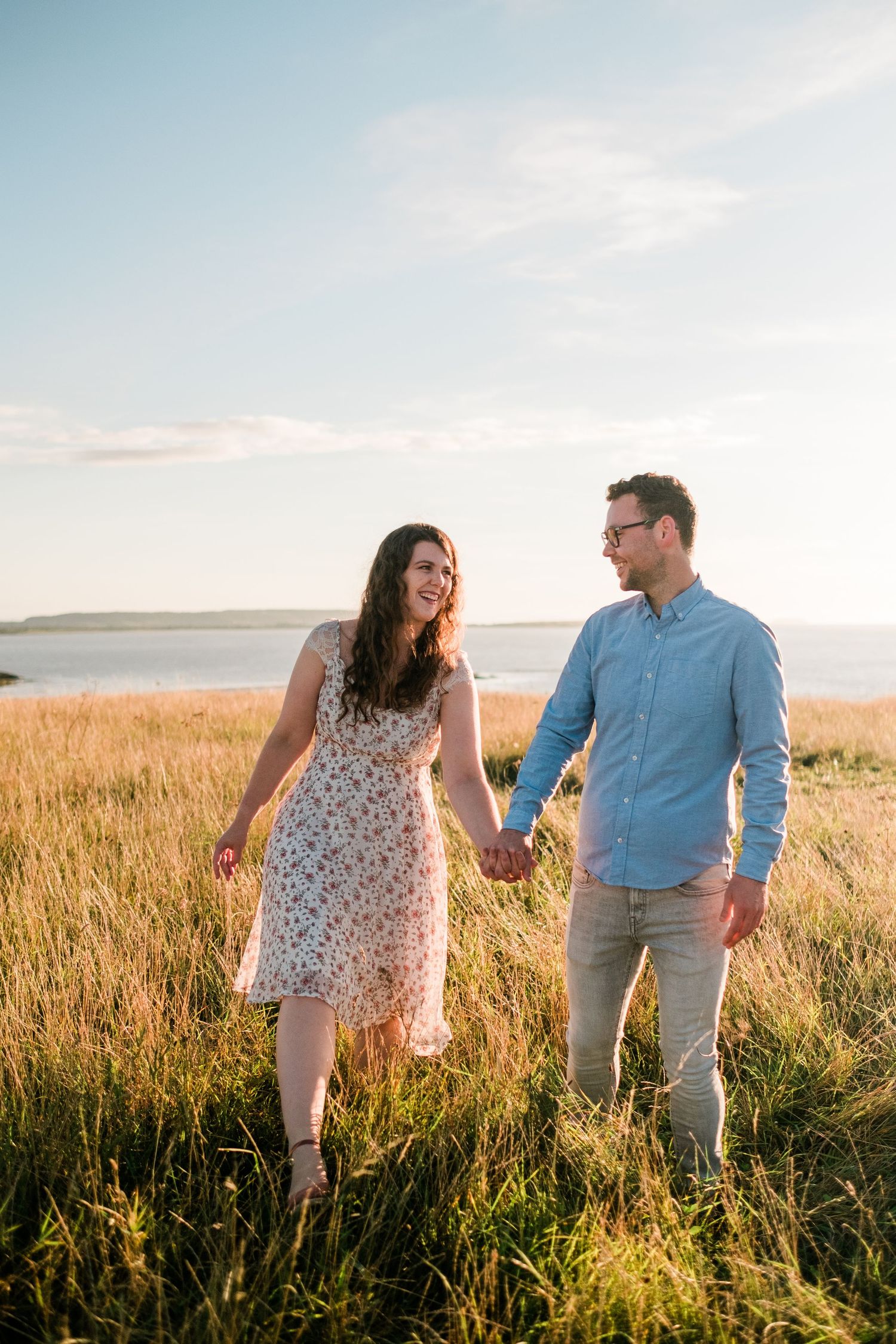 A couple strolls through tall grass in a coastal meadow during golden hour.