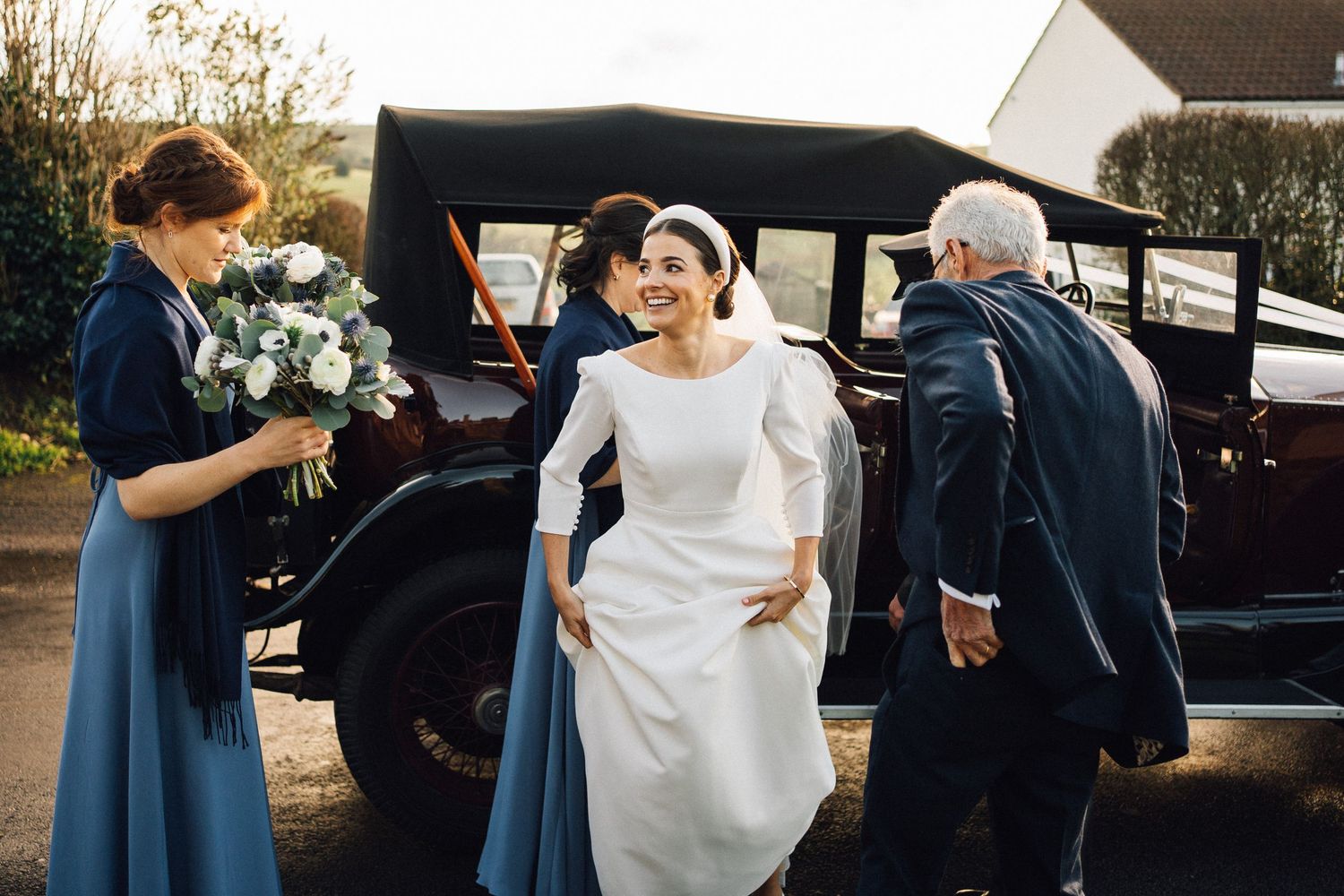 Wedding party members in navy blue dresses help the bride into a car on a sunny afternoon.