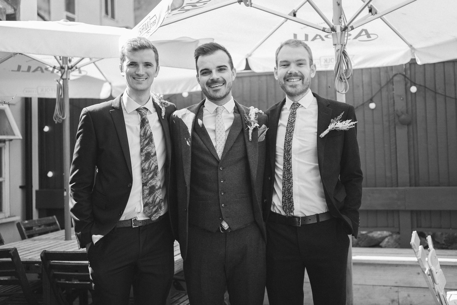 Groomsmen in formal suits pose together outdoors for wedding party photos.