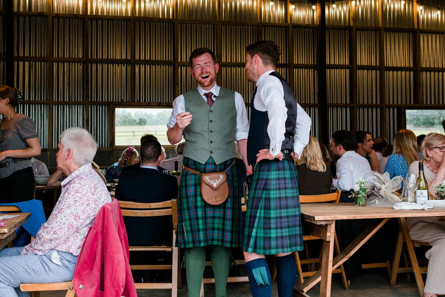 Wedding guests enjoying a reception dinner in a rustic barn setting with ambient lighting.