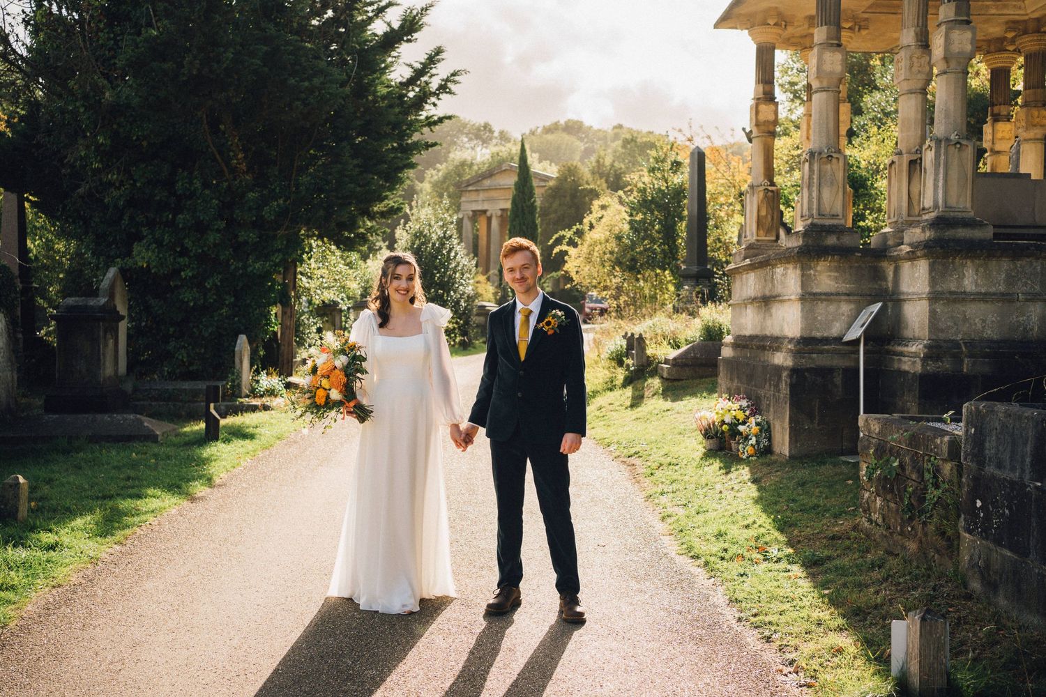 A couple in wedding attire walks along a scenic path surrounded by greenery and stone architecture.