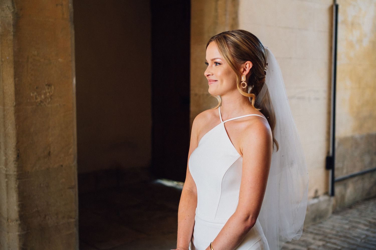 A bride in a sleeveless white wedding gown and veil smiles while standing in a stone archway.