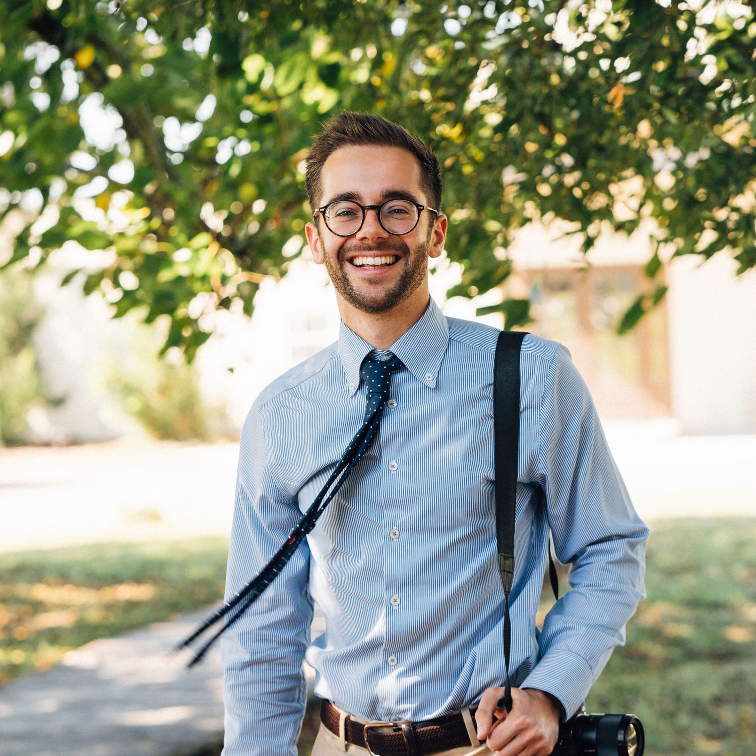 Smiling young man in glasses and blue shirt with tie holding camera outdoors under green trees.